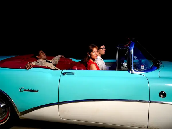 Turquoise and white vintage convertible car at night, with two people sitting inside.