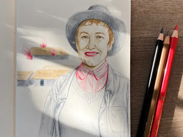 Coloured pencil portrait of woman with grey hat, pink collar, and white cardigan on wooden surface beside black, beige, and red pencils.