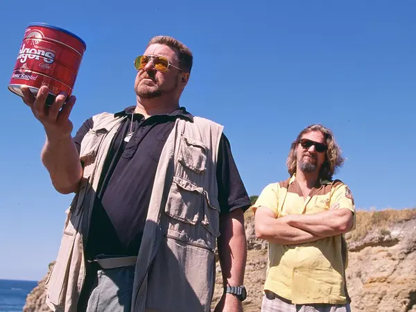 Two men standing in a desert landscape, one holding a red container while the other has his arms crossed.