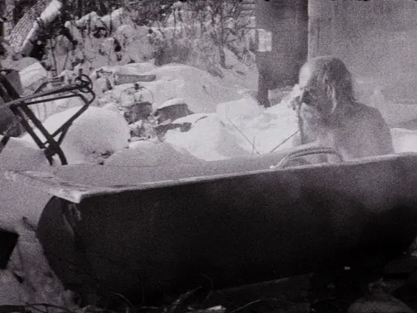 Black and white image of a person lying in a bed, surrounded by snow-covered furniture and decor.