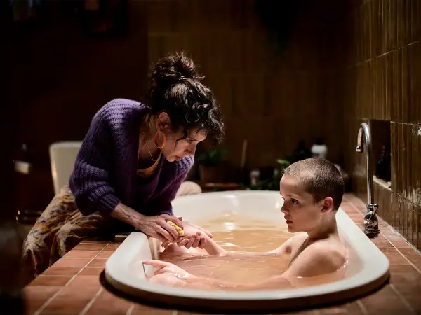 Woman in purple jumper leaning over white bathtub, assisting person with short hair bathing in warm-lit bathroom setting.