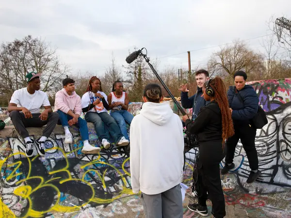 A group of young people sitting on benches, with one person speaking into a microphone in front of them. The scene is set against a backdrop of colourful graffiti-covered walls.