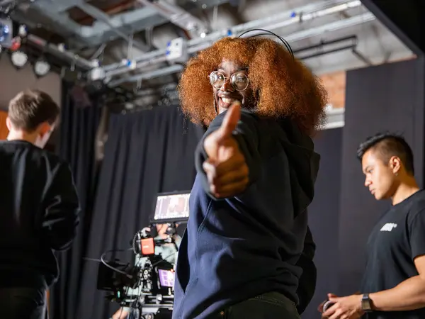 Person with curly orange hair wearing glasses and a black outfit, sitting on a chair on a stage.
