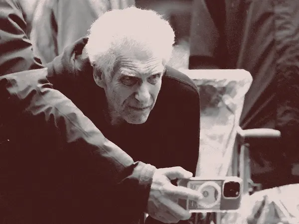 An elderly man with white hair and a stern expression, sitting in a chair and holding a camera.