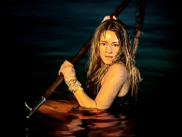 A young woman in the water, holding a wooden pole, with a warm-toned lighting creating a dramatic atmosphere.