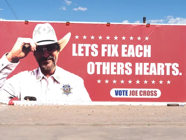 Red billboard with white text "LETS FIX EACH OTHERS HEARTS. VOTE JOE CROSS" featuring man in cowboy hat and uniform against blue sky.