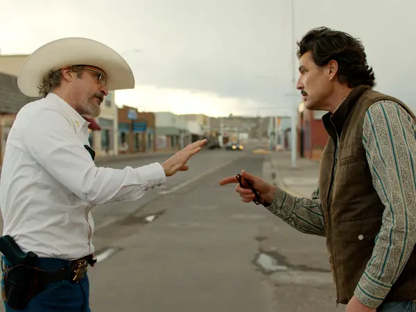 Two men in cowboy hats facing each other on a dusty road with buildings in the background.