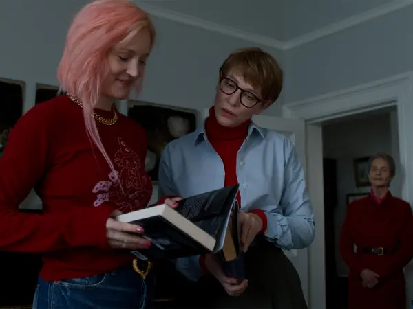 Woman with pink hair in red jacket and man in glasses and light blue shirt examining a black book together indoors.