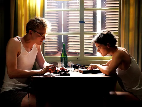 Two people playing sitting at a table, bottle of drink on the table, warm lighting from window.