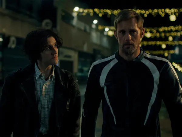 Two men standing outdoors at night, one in a jacket, the other in a casual shirt, under twinkling lights.