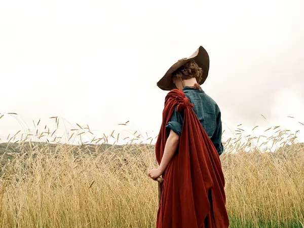 Person in dark hat, blue top, and red fabric wrap standing in golden grassland with birds flying overhead against pale sky.