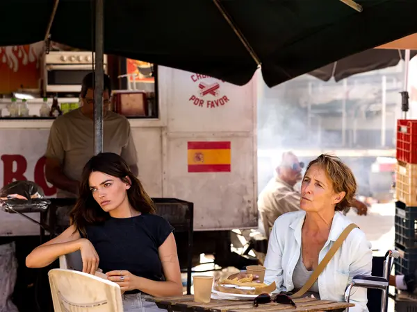 Two women sitting at outdoor café table under umbrellas, one in dark top, one in white shirt, Spanish flag visible on wall behind.