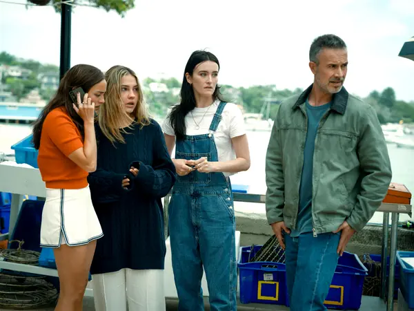 Four people standing on boat deck: woman in orange top on phone, blonde woman in navy jumper, brunette in denim dungarees, man in grey jacket.