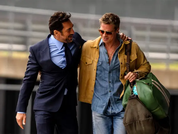 Two men walking together, one wearing a navy suit and the other in a yellow jacket and jeans.