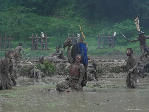 Samurai in traditional uniform, covered in mud, standing in a muddy river with trees and foliage in the background. One of them holds a human head aloft.