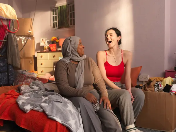 Two women sitting on a bed surrounded by clutter and personal items.