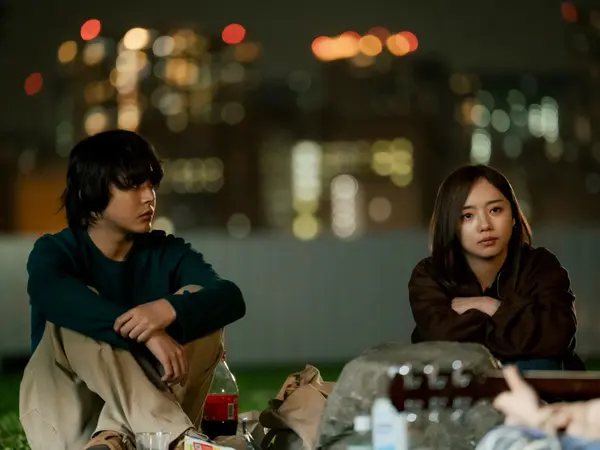 Two people sitting on a bench at night, surrounded by blurred city lights in the background.