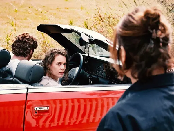 Two individuals, possibly actors, seated in a red convertible car against a rural backdrop.