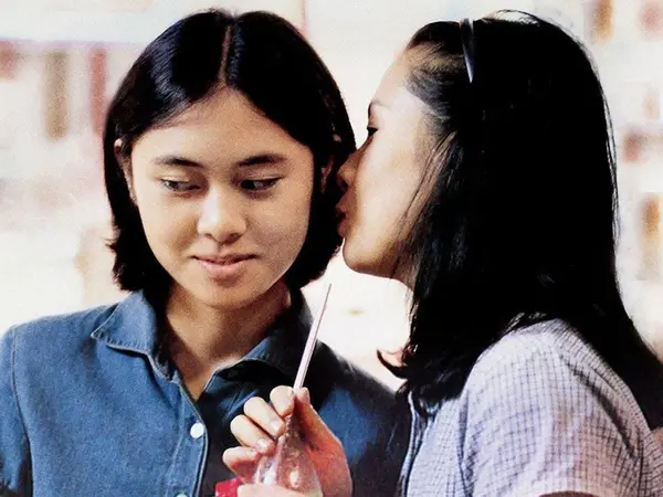 Two young Chinese women stand together, one in a blue denim shirt, the other in a purple and white gingham shirt. The one in the purple and white shirt is holding a soda bottle with a straw, and is whispering in the other girl's ear.