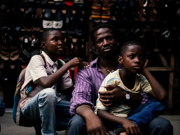 Three African men seated in a dimly lit stall, wearing casual clothing. The men appear to be in conversation, with the younger ones looking up at the older man.