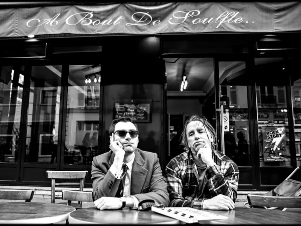 Exterior of a cafe, two men sitting at a table in the foreground, one wearing a suit and the other a checkered shirt, text 'A bout de Souffle' on the storefront awning.