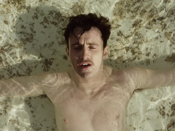 A man with dark hair and a pained expression on his face, his upper body visible against a mottled green and grey background.