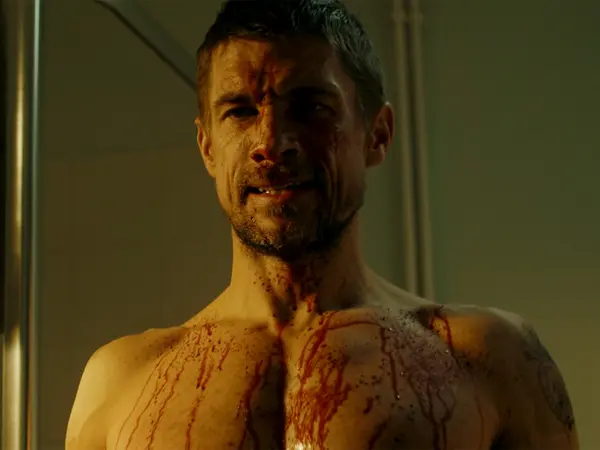 A close-up of a shirtless man with a bloodied and bruised face, his expression stern and intense.