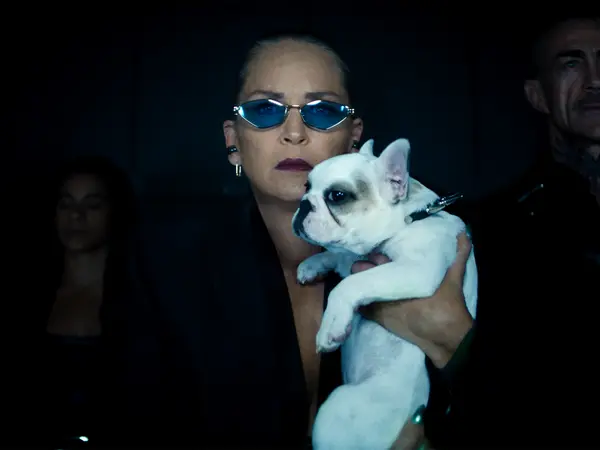 Woman wearing blue sunglasses holds white French bulldog in dark setting with people in background.