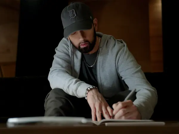 Man in grey baseball cap and hoodie sits at table, looking down whilst writing or reading documents in dimly lit room.
