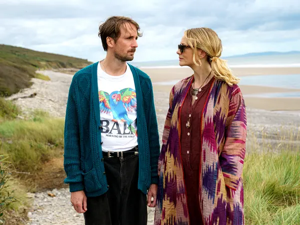 Two people, a man and a woman, standing on a beach with grassy hills in the background. The man is wearing a colourful graphic T-shirt and a teal cardigan, while the woman is wearing a patterned, multi-coloured coat.
