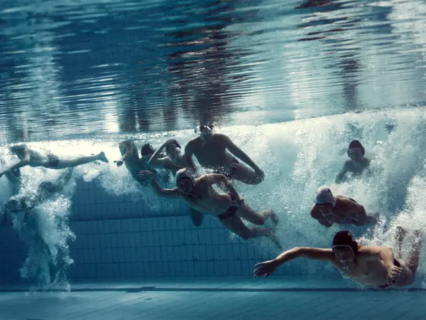Several swimmers underwater in a pool, creating splashes and distorted reflections in the water.