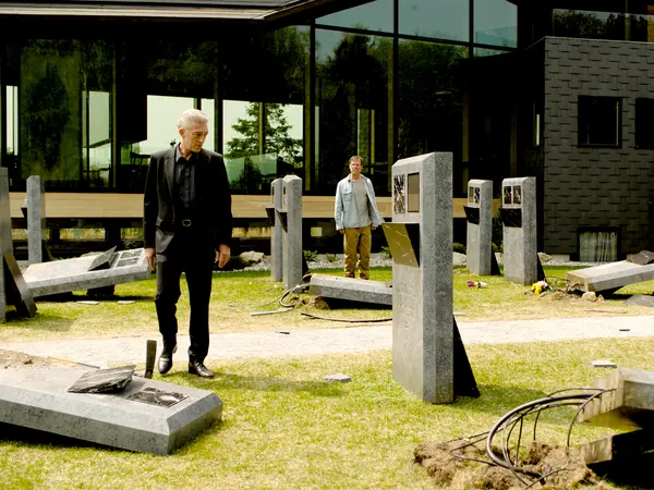 Two men standing amongst concrete construction materials and metal posts on grass with modern glass building in background.