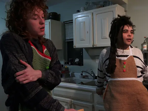 Two people, one wearing a black jacket and the other wearing a brown checked dress, standing in a kitchen.