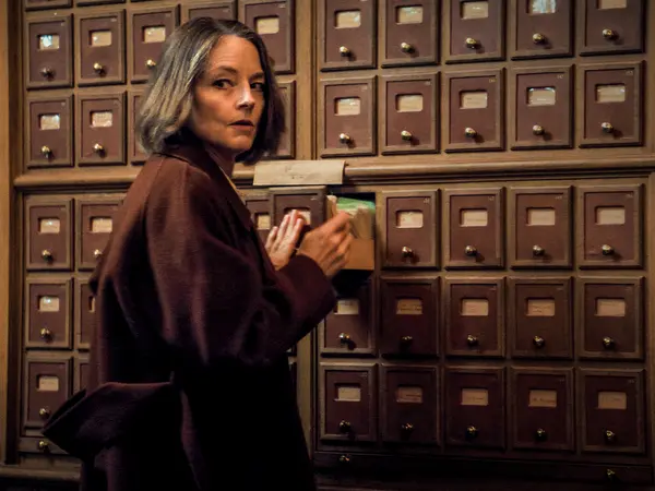 Woman in brown coat stands in front of wall of wooden drawers.