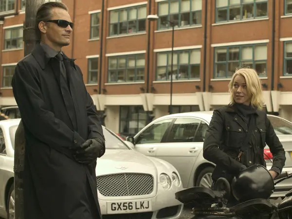 Two individuals, a man and a woman, standing in front of a parked Bentley motor car on a city street.