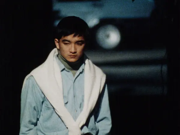 A young East Asian man wearing a blue shire with a white sweater around his shoulders stands on a street with an uncertain expression.