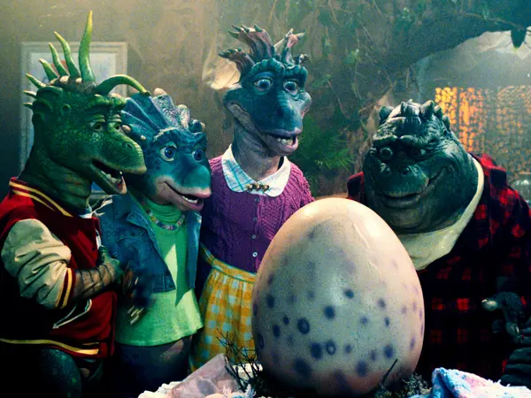 Three fantasy creatures wearing colourful, patterned clothing surrounding a large spotted egg on a plush surface.