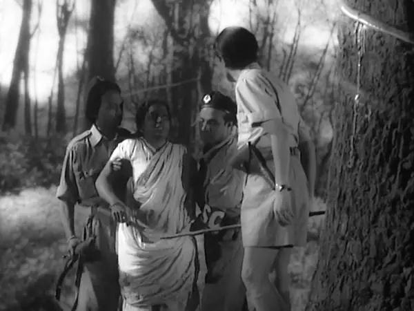 A black an dwhite image of four people standing by a tree in a dark forest. Two men are holding a woman, while another has his back to the camera looking down at her.